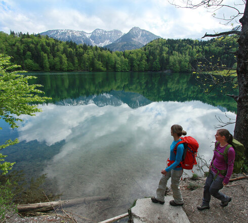 Das Bild zeigt zwei Frauen, die Wanderkleidung und Rucksäcke tragen. Sie stehen am Ufer eines Sees. Die Wasseroberfläche des Sees ist ganz ruhig. Der See ist mit hohen Bäumen eingerahmt. Im Hintergrund erheben sich die Berge. Der Himmel ist blau. Wolken ziehen auf.   