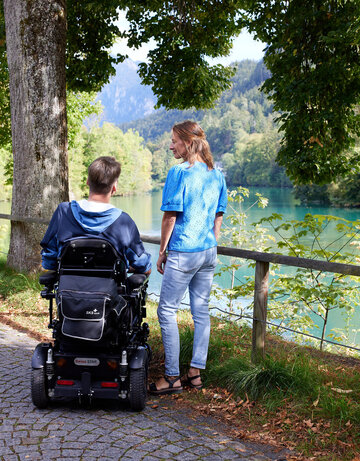Das Bild zeigt einen Mann und eine Frau. Der Mann sitzt in einem schwarzen Rollstuhl, während die Frau neben ihm steht. Beide blicken in dieselbe Richtung, ihr Blick scheint auf das friedliche Flussufer im Hintergrund gerichtet zu sein. Der Fluss Lech, dessen Wasser ein schönes Türkis aufweist, schlängelt sich durch üppige Grünanlagen. Bäume und Sträucher säumen das Bild. Der Weg, auf dem die beiden stehen, wird von einer Holzumzäunung gesäumt.