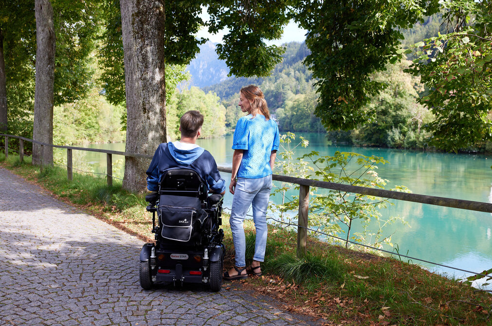 Das Bild zeigt einen Mann und eine Frau. Der Mann sitzt in einem schwarzen Rollstuhl, während die Frau neben ihm steht. Beide blicken in dieselbe Richtung, ihr Blick scheint auf das friedliche Flussufer im Hintergrund gerichtet zu sein. Der Fluss Lech, dessen Wasser ein schönes Türkis aufweist, schlängelt sich durch üppige Grünanlagen. Bäume und Sträucher säumen das Bild. Der Weg, auf dem die beiden stehen, wird von einer Holzumzäunung gesäumt.