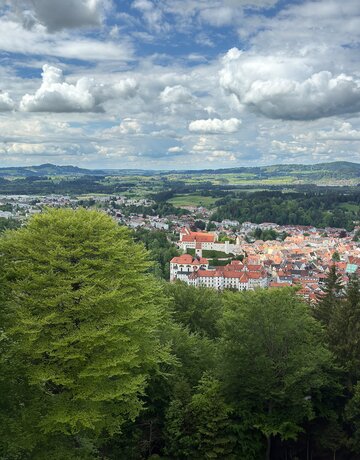 Das Bild zeigt Füssen von oben. Es ist von einer Anhöhe aus aufgenommen. Über grüne Baumwipfel hinweg erblickt man die roten Dächer der Stadt. Im Vordergrund sind die großen Gebäudekomplexe des ehemaligen Benediktinerklosters St. Mang und des Hohen Schlosses zu sehen. Im Hintergrund erheben sich bewaldete Hügel. 