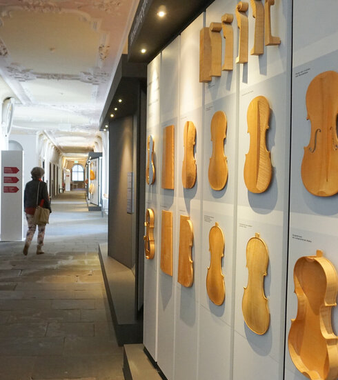 Man blickt einen langen Gang entlang. Die Decke ist reich mit Stuck verziert. Am Boden sind Steinplatten verlegt. Auf der linken Seite des Ganges führen Türen in die dahinterliegenden Zimmer. Über den Türen befinden sich mit Stuck eingerahmte Bilder. Einige Stuckrahmen sind leer. Auf der rechten Seite befinden sich zwei Schaukästen, die die unterschiedlichen Entstehungsstadien einer Geige präsentieren. 
