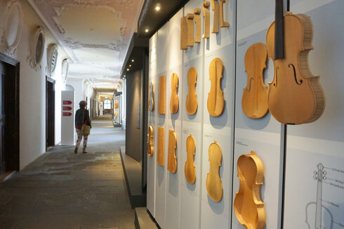 Man blickt einen langen Gang entlang. Die Decke ist reich mit Stuck verziert. Am Boden sind Steinplatten verlegt. Auf der linken Seite des Ganges führen Türen in die dahinterliegenden Zimmer. Über den Türen befinden sich mit Stuck eingerahmte Bilder. Einige Stuckrahmen sind leer. Auf der rechten Seite befinden sich zwei Schaukästen, die die unterschiedlichen Entstehungsstadien einer Geige präsentieren. 
