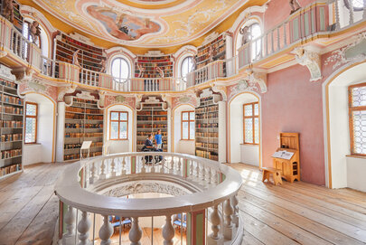 Das Bild zeigt eine imposante, kreisrunde Bibliothek mit einer hohen Decke. Die Wände sind in einem warmen, rosafarbenen Ton gemalt, und die Decke ist mit aufwendigen Mustern und wunderschönen Verzierungen geschmückt.