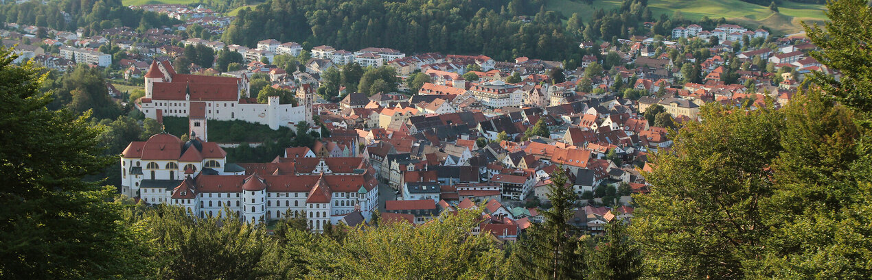 Das Bild zeigt Füssen von oben. Es ist von einer Anhöhe aus aufgenommen. Über grüne Baumwipfel hinweg erblickt man die roten Dächer der Stadt. Im Vordergrund sind die großen Gebäudekomplexe des ehemaligen Benediktinerklosters St. Mang und des Hohen Schlosses zu sehen. Im Hintergrund erheben sich bewaldete Hügel. Davor liegt ein blauer See.