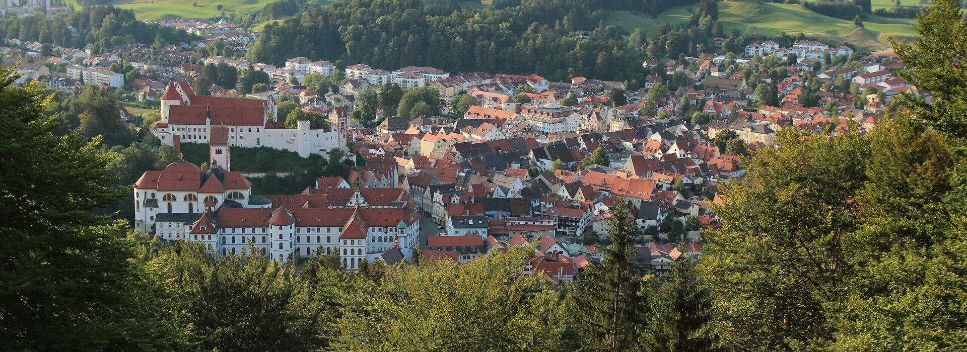 Das Bild zeigt Füssen von oben. Es ist von einer Anhöhe aus aufgenommen. Über grüne Baumwipfel hinweg erblickt man die roten Dächer der Stadt. Im Vordergrund sind die großen Gebäudekomplexe des ehemaligen Benediktinerklosters St. Mang und des Hohen Schlosses zu sehen. Im Hintergrund erheben sich bewaldete Hügel. Davor liegt ein blauer See.