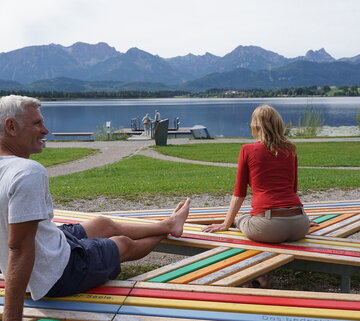 Das Bild zeigt einen Mann und eine Frau, die auf einer bunten Holzbank sitzen. Die Holzbank besteht aus mehreren Latten, die kreuz und quer miteinander verschraubt sind. In die bunten Latten sind Sätze eingraviert, die aus der aufgenommenen Perspektive nicht lesbar sind. Der Mann streckt seine Beine auf die Bank und stützt sich nach hinten auf seine Arme. Vor der Bank ist die Uferpromenade zu sehen. Im Wasser in unmittelbarer Ufernähe schwimmt eine hölzerne Insel. Die Wasseroberfläche des Sees ist ruhig. Im Hintergrund erheben sich die Berge. Der Himmel ist leicht bedeckt.  