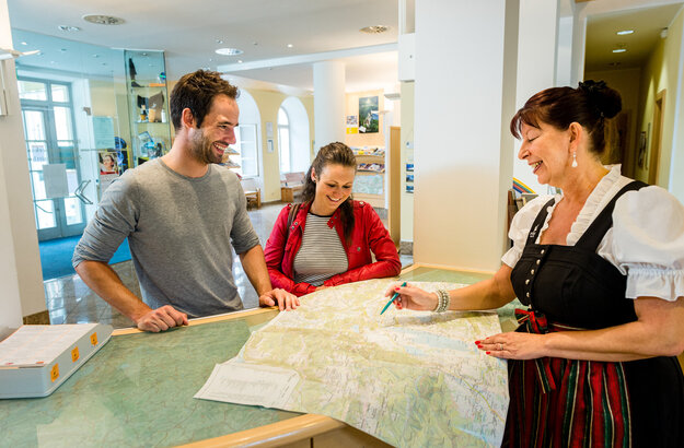 Das Bild zeigt einen Mann und zwei Frauen, die an einem Informationstresen stehen. Der Mann und die Frau, die hinter dem Tresen stehen, tragen Freizeitkleidung. Die Dame auf der anderen Seite trägt ein Dirndl mit einer grün-roten Schürze. Alle lächeln. Die Frau im Dirndl deutet mit einem Stift auf eine Karte, die vor den Personen auf dem Tresen liegt. Alle lächeln.