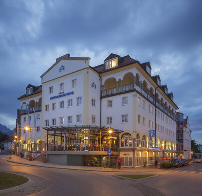  Das Bild zeigt ein großes, mehrstöckiges Gebäude mit einer markanten weißen Fassade. Das Gebäude zeigt eine Mischung aus architektonischen Stilen, einschließlich eines zentralen Turms mit Kuppel und einer Reihe von Bogenfenstern. Das Dach des Gebäudes ist mit mehreren Schornsteinen verziert. Das Gebäude wird von zahlreichen Lichtern beleuchtet, die seine Fassade und die umliegende Fläche hervorheben. Vorne im Vordergrund ist eine Straße zu sehen, auf der einige Autos am Straßenrand geparkt sind. Der Himmel über dem Bild ist wolkig, was auf den Anbruch des Tages oder auf den Einbruch der Dämmerung hindeutet. Die Gesammtatmosphäre des Bildes wirkt friedlich und einladend.