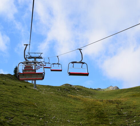 Das Bild zeigt eine Sesselbahn im Sommer. Darunter liegt ein Hang auf dem grünes Gras wächst. Der Himmel ist blau mit weißen Wolken. Die Sonne scheint.  