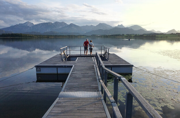 Das Bild zeigt einen großen Steg, der in den Hopfensee ragt. Auf dem See schwimmen Seerosenblätter. Am Ende des Stegs wird öffnet sich eine Plattform, in deren Mitte ein Kneipptretbecken eingelassen ist. Auf der Kneippinsel stehen zwei Menschen mit dem Rücken an ein Geländer gelehnt und blicken in die Ferne. Der Himmel ist in ein ruhiges Abendlicht getaucht und im Hintergrund erhebt sich die Bergkette der Allgäuer Alpen.