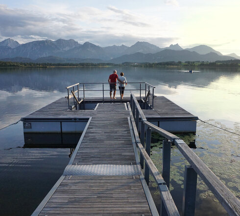 Das Bild zeigt einen großen Steg, der in den Hopfensee ragt. Auf dem See schwimmen Seerosenblätter. Am Ende des Stegs wird öffnet sich eine Plattform, in deren Mitte ein Kneipptretbecken eingelassen ist. Auf der Kneippinsel stehen zwei Menschen mit dem Rücken an ein Geländer gelehnt und blicken in die Ferne. Der Himmel ist in ein ruhiges Abendlicht getaucht und im Hintergrund erhebt sich die Bergkette der Allgäuer Alpen.