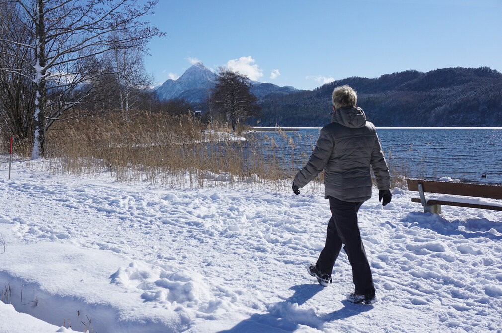 Das Bild erfasst eine ruhige Winterlandschaft. Eine Person, in einem grauen Mantel und schwarzen Hosen gekleidet, geht auf einem schneebedeckten Weg. Der Weg wird von Bäumen und Büschen flankiert, deren Äste unter der Last des Schnees hängen. Die Person geht am Ufer des gefrorenen Weissensees entlang. In der Ferne erheben sich majestätische Berge gegen den Hintergrund eines klaren blauen Himmels. 