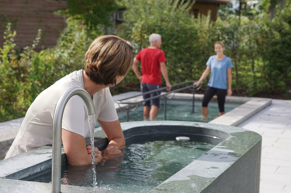 Das Bild zeigt im Vordergrund eine Frau, die ihre unbekleideten Unterarme in ein Armbecken taucht. Aus einem Wasserhahn davor läuft Wasser in das Armbecken. Im Hintergrund sieht man einen Mann und eine Frau in einem Wassertretbecken stehen. Die Wasserbecken sind mit Büschen und Bäumen im Hintergrund eingerahmt. Die Sonne scheint. 