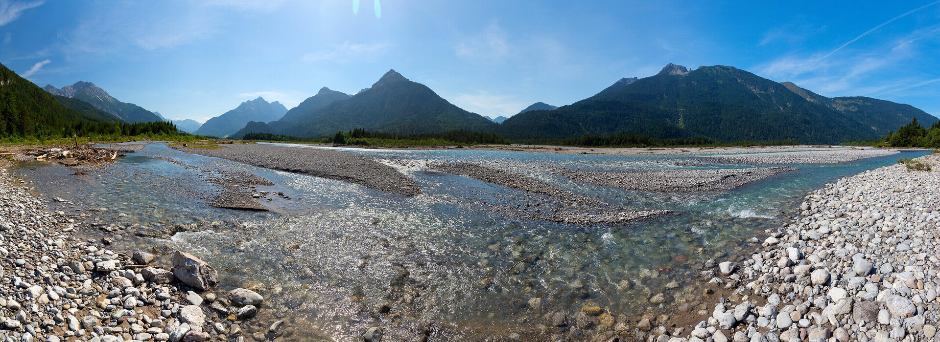 Auf dem Bild sieht man ein Flussbett umrahmt von Wald. Im Hintergrund sind Berge zu erkennen. Sie Sonne strahlt über das Bild.