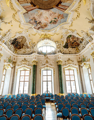 Das Foto zeigt eine prunkvollen Raum. Die Decke ist reich mit Stuck und mehreren Deckengemälden verziert. Den Raum säumen hohe Fenster. Auch die Fensterrahmen sind mit weiß, goldenem Stuck verziert. Der Raum wird von vier Säulen eingerahmt. Zwei Säulen sind Lapislazuli blau, zwei malachitgrün. Der Raum steht voller Holzstühle, die mit blauem Stoff bezogen sind. Die Stühle sind alle Richtung Kamera ausgerichtet. Ein schmaler Gang führt durch die Stühle hindurch. Der Raum ist lichtdurchflutet.  