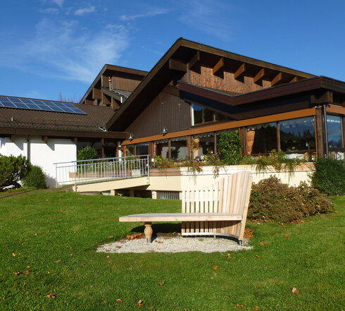  Das Bild zeigt eine idyllische Außenansicht. Im Zentrum der Szene steht eine hölzerne Bank, die Passanten einlädt, Platz zu nehmen und die Aussicht zu genießen. Die Bank steht auf einem üppigen grünen Rasen, der von heruntergefallenen Blättern gepunktet ist, was auf die sich verändernden Jahreszeiten hinweist.  Im Hintergrund erhebt sich ein großes hölzernes Gebäude. Sein rustikaler Charme wird durch eine Solarzelle auf dem Dach betont, die auf einen umweltfreundlichen Ansatz des Energieverbrauchs hindeutet. Die Architektur des Gebäudes wird durch eine Veranda ergänzt, die mit Kübelpflanzen geschmückt ist, was der Struktur einen Hauch von Natur verleiht.  Der Himmel darüber ist ein hellblau, durchzogen von flauschigen weißen Wolken, die eine friedliche und idyllische Atmosphäre erschaffen. Die Gesamtansicht ist eine der Ruhe und Harmonie mit der Natur.