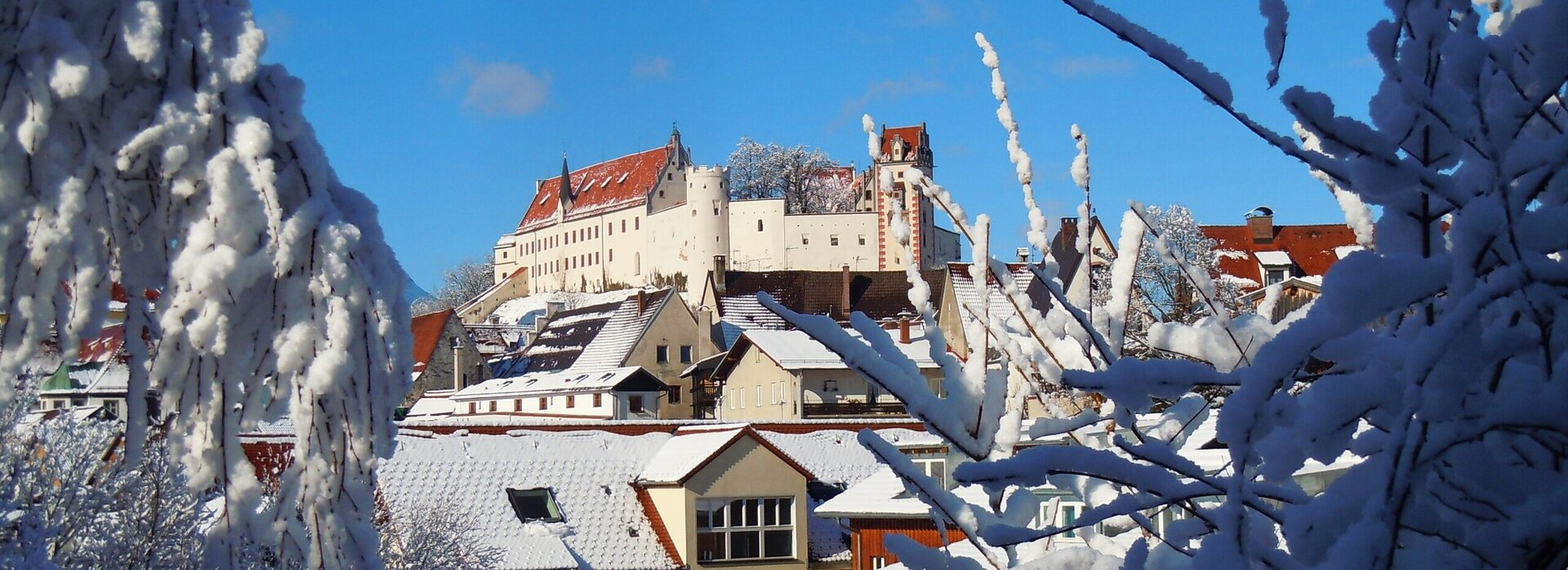 Auf dem Bild ist durch Baunzweige hindurch eine verschneite Ansicht von Füssen und dem Hohen Schloss im Hintergrund zu sehen. Die Dächer sind mit Schnee bedeckt und vermitteln eine winterliche, schöne Atmosphäreige Gebäude auch.