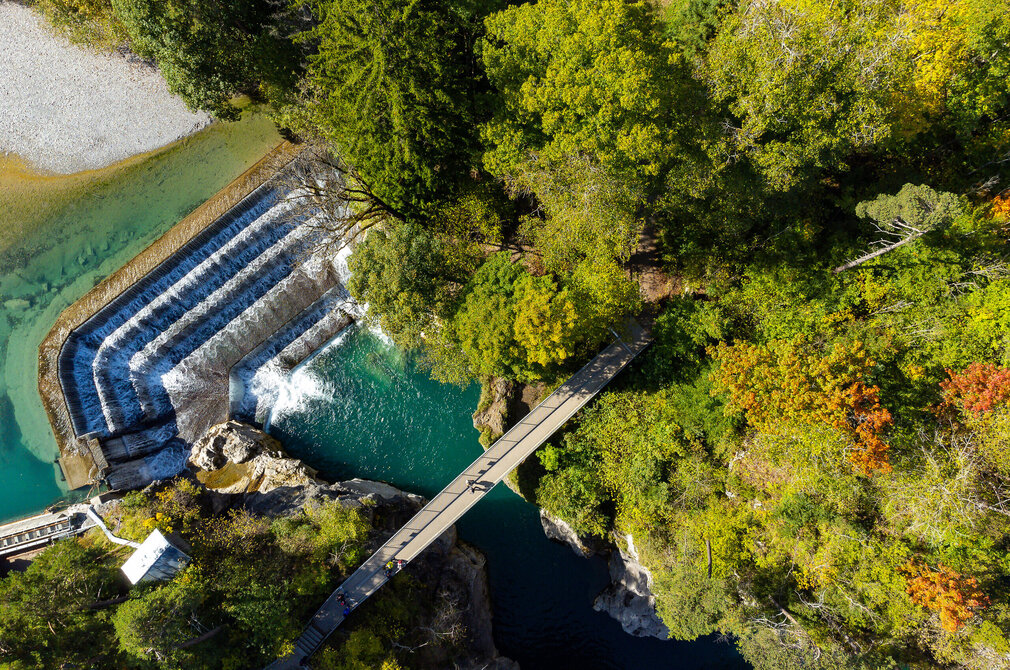 Das Bild zeigt eine Luftaufnahme einer friedlichen Landschaft, die vom Fluss Lech dominiert wird, der sich durch die Szene windet. Eine Brücke überspannt den türkisen Fluss und stellt eine Verbindung zwischen zwei ungesehenen Punkten her. Das Ufer wird gesäumt von Bäumen und Sträuchern, deren Laub sich an einigen Stellen bereits rot und gelb färbt und so auf den beginnenden Herbst hindeutet.