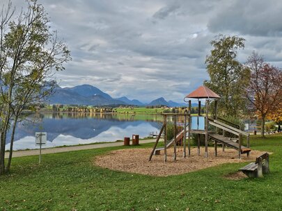 Auf dem Bild sieht man einen Spielplatz mit einem Rutscheturm. Im Hintergrund ist ein See zu sehen, der vor einer Bergkulisse liegt.