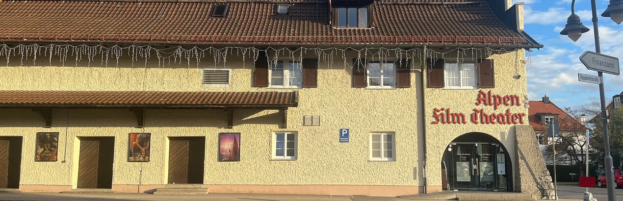 Das Bild zeigt ein Gebäude mit einer beigefarbenen Fassade und einem braunen Dach. Über der Eingangstüre steht in roter Schrift Alpenfilmtheater.