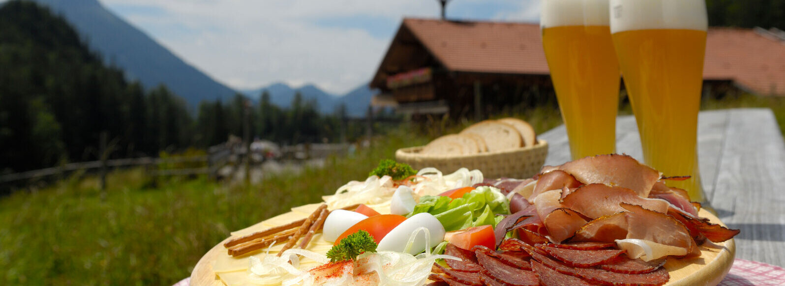 Das Bild zeigt einen hölzernen Picknicktisch mit einem Teller Essen und zwei Bieren. Der Tisch ist in einer schönen Außenkulisse aufgestellt, mit einem Berg im Hintergrund.