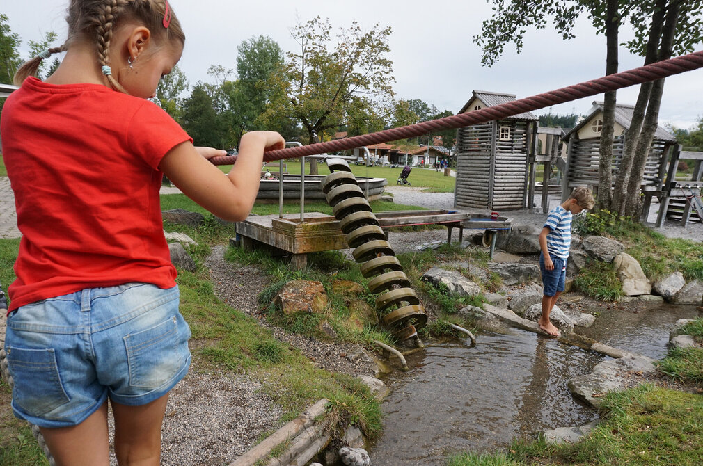 Das Bild zeigt eine lebhafte Szene auf einem Spielplatz. Der Hauptfokus liegt auf einem jungen Mädchen, das in einem leuchtend roten Hemd und blauer Jeans gekleidet ist und aktiv mit einem großen, roten Seil spielt. Sie steht auf einem grünen Rasen, ihre ganze Aufmerksamkeit ist auf das Seil gerichtet. Im Hintergrund ist ein Junge zu sehen. Er steht auf einem felsigen Bereich, seinen Blick auf die Mädchen gerichtet und das Seil. Der Spielplatz selbst ist eine Mischung aus natürlichen und von Menschen geschaffenen Elementen. Der grüne Rasen, auf dem das Mädchen spielt, ist üppig und grün, bietet eine weiche und sichere Fläche für ihr Spiel. 