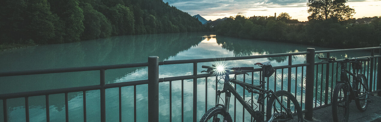Das Bild zeigt im Vordergrund ein Brückengeländer, an dem zwei Mountainbikes lehnen. Unter der Brücke fließt ruhig der Fluß Lech. An dessen Ufer wachsen üppige grüne Wälder. Die Sonne geht gerade unter. Am Himmel ziehen Wolken.  