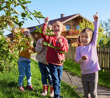 Das Bild zeigt eine herrliche Szene von drei Kindern in einem Garten. Der Garten ist reich an grünen Pflanzen und hat einen hölzernen Zaun im Hintergrund. Die Kinder stehen vor dem Zaun und ihre Gesichter sind erfüllt von Freude.  Das Kind auf der linken Seite hält einen Ast mit grünen Blättern, als wäre es ein geschätzter Besitz. Das mittlere Kind hält eine Blume, vielleicht ein Lieblingsblüher oder eine neue Entdeckung. Das Kind auf der rechten Seite zeigt nach oben, möglicherweise auf einen Vogel oder einen Baum.  Ihre Gesichtsausdrücke und Handlungen suggerieren ein Gefühl von Wunder und Erkundung. Der Garten, mit seiner lebhaften Grünfläche und dem hölzernen Zaun, bietet eine ruhige und sichere Umgebung für die Kinder, um zu spielen und zu lernen. Das Bild bildet einen Moment von Kindheitsunschuld und Neugierde schön ab.
