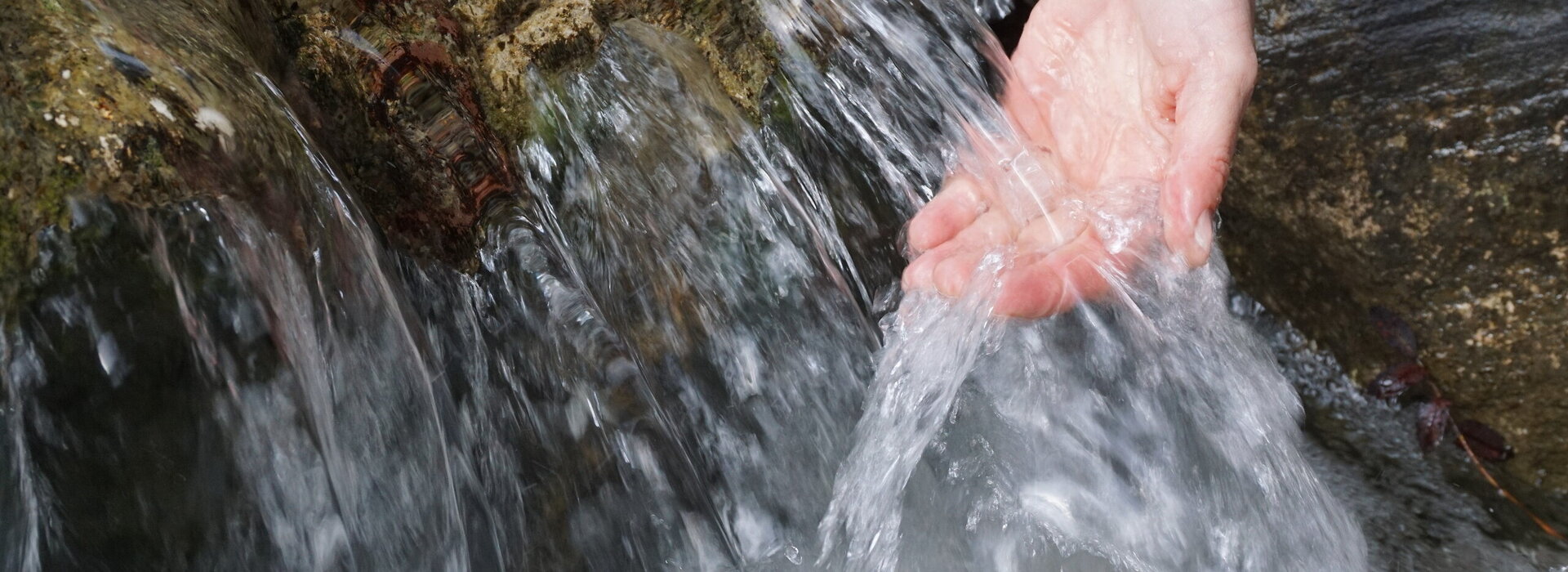 Das Bild zeigt eine Nahaufnahme der Hand einer Person, die unter einem Wasserlauf gehalten wird. Das Wasser aus dem Bach fließt über einen felsigen Absatz in einem kleinen Wasserfall sprudelnd nach unten. 