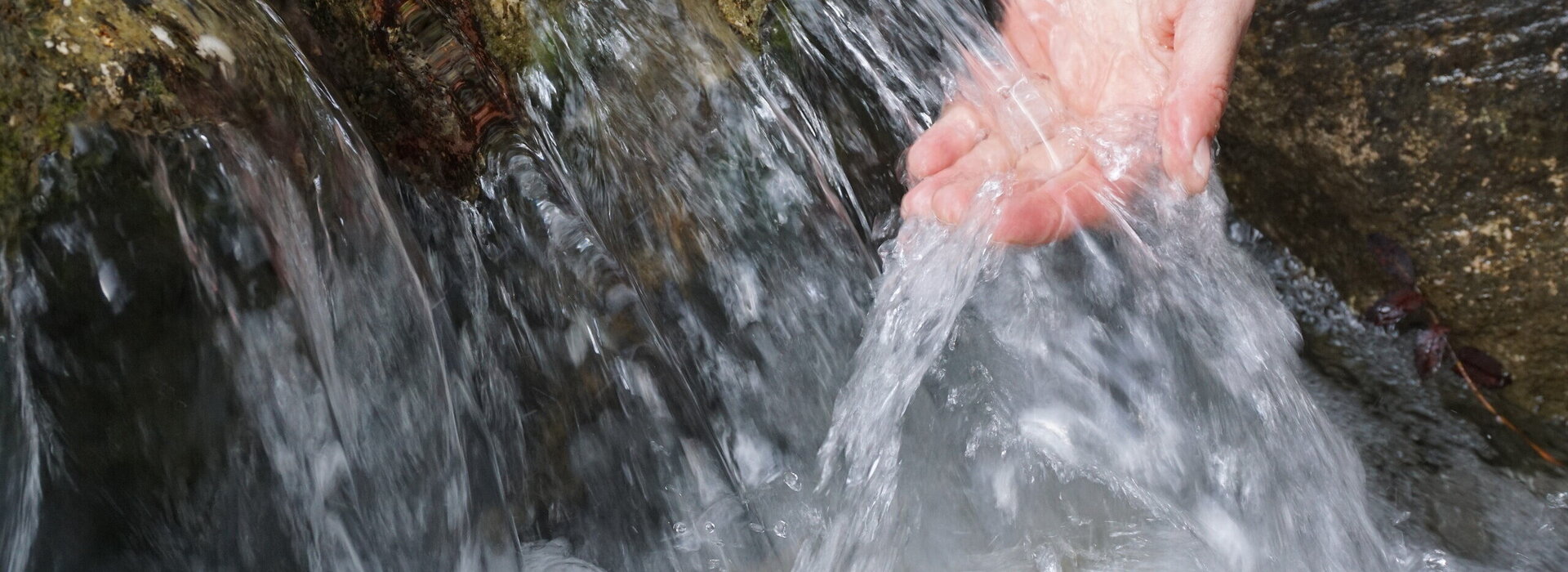 Das Bild zeigt eine Nahaufnahme der Hand einer Person, die unter einem Wasserlauf gehalten wird. Das Wasser aus dem Bach fließt über einen felsigen Absatz in einem kleinen Wasserfall sprudelnd nach unten. 