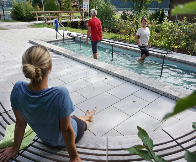 Das Bild zeigt zwei Personen, die in einem Tretbecken Wasser treten. Sie haben die Hosenbeine hochgekrempelt. Eine Person blickt auf eine Frau, die auf einer halbrunden Holzbank neben dem Tretbecken sitzt. Diese Frau hat dem Betrachter den Rücken zugedreht. Die Sonne scheint. 