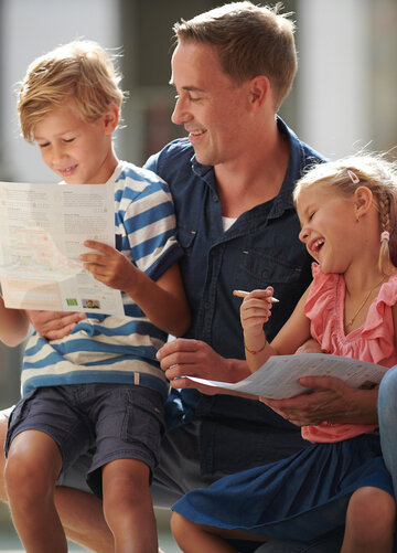 Das Bild ist ein Farbfoto, das eine Familie von vier Personen zeigt, darunter zwei Erwachsene und zwei Kinder, die zusammen auf einer Parkbank sitzen. Der Mann sitzt auf der linken Seite der Bank, die Frau auf der rechten. Zwischen ihnen sitzen zwei Kinder, ein Junge und ein Mädchen. Der Mann trägt ein blaues Hemd und strahlt in die Kamera. Die Frau trägt ein pinkfarbenes Hemd und blickt ebenfalls lächelnd in die Kamera. Die Kinder beschäftigen sich gerade mit einer Aktivität, das Mädchen hält ein Buch in der Hand, der Junge schaut auf ein Stück Papier. Die Familie scheint, sich in dem Moment, in dem das Foto aufgenommen wurde, zu freuen. Der Hintergrund ist verschwommen, aber er vermittelt den Eindruck einer städtischen Umgebung mit Gebäuden und einem Baum.