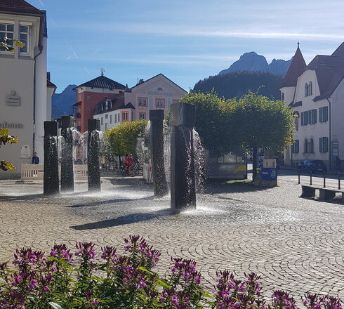 In diesem Bild wird eine friedliche Außenlandschaft eingefangen, die einen Springbrunnen in den Mittelpunkt stellt. Rund um den Springbrunnen befinden sich verschiedene Pflanzen, darunter lila Blumen und grüne Sträucher, was der Szene einen Hauch von Natur verleiht. Der Springbrunnen liegt auf einem gepflasterten Weg, der links von einem weißen Gebäude und rechts von einem gelben Gebäude flankiert wird. Der Himmel ist ein tiefes Blau, was auf einen angenehmen Tag hinweist. Die allgemeine Atmosphäre des Bildes ist ruhig und einladend, was den Ort zum Entspannen und Nachdenken hervorragend macht.