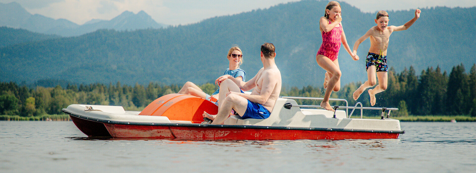 Das Bild zeigt ein Tretboot, das auf einem See schwimmt. An den Tretpositionen des Boots sitzen ein Mann und eine Frau. Der Mann trägt Badekleidung, die Frau eine kurze Hose und ein T-Shirt. Im hinteren Bereich des Tretboots springen ein Junge und ein Mädchen in Badekleidung ins Wasser. Das Mädchen hält sich die Nase zu. Im Hintergrund erheben sich die bewaldeten Berge. Der blaue Himmel ist von Wolken durch zogen.   