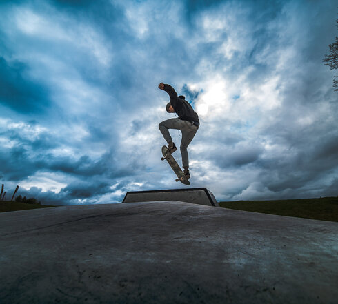  Das Bild fängt eine spannende Szene in einem Vergnügungspark ein. Der Fokus liegt auf einer großen, bunten Struktur, die einer Kletterwand ähnelt. Diese Struktur besteht aus verschiedenen Holzpfosten und Plattformen, die durch eine Vielzahl von Seilen und Drähten miteinander verbunden sind.  Mehrere Menschen sind dabei zu sehen, wie sie mit dieser Struktur interagieren, einige von ihnen klettern die Pfosten hinauf, während andere von den Plattformen schwingen. Die Struktur ist nicht nur ein Spielplatz, sondern auch ein Ort für Abenteuer.  Im Vordergrund gibt es eine kleine, hölzerne Plattform mit einer Brüstung. Diese Plattform ist mit einer Statue eines Ziegenbocks verziert, was der Szene eine Prise Fantasie verleiht.  Der Himmel darüber ist ein klares Blau, durchzogen von weißen Wolken, und bildet einen wunderschönen Hintergrund für die bunte Struktur. Das Bild ist eine lebendige Darstellung von Spaß und Abenteuer in einem Vergnügungspark.