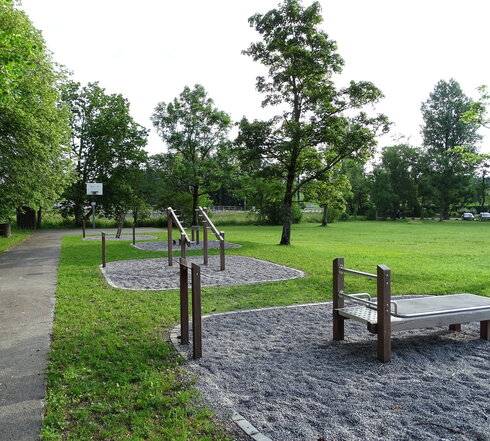 Das Bild zeigt eine Wiese, die durch mehrere Runde Kiesfelder unterbrochen ist. Auf den abgerundeten Kiesfeldern stehen unterschiedliche Sportgeräte aus Holz und Metall. An jedem Sportgerät ist eine Tafel mit der jeweiligen Anleitung angebracht. Im Hintergrund ist ein Basketballfeld über einem asphaltierten Spielfeld angebracht. Der Bereich ist mit hohen, grünen Bäumen eingerahmt. Der Himmel ist bedeckt. 