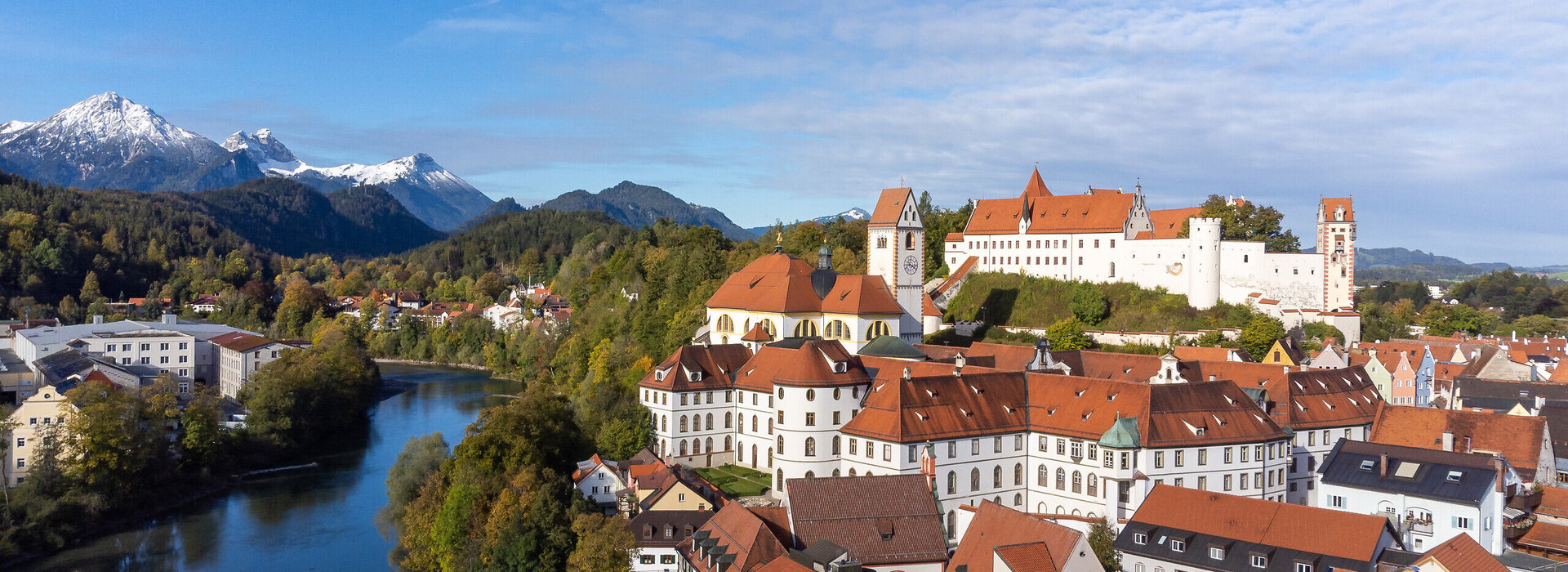 Das Foto zeigt das Hohen Schloss, dass auf einer Anhöhe über der Altstadt von Füssen thront. Direkt darunter ist der große Klosterkomplex des ehemaligen Benediktinerklosters mit seinen vielen Fenstern zu sehen. Alle Gebäude haben rote Dächer und weiße Wände. Neben den Gebäuden fließt der Lech, über den einen Brücke führt. Im Hintergrund erheben sich die Alpen, deren Gipfel schneebedeckt sind. Die Bäume haben bunte Blätter. Der Himmel ist ein wenig wolkenverhangen. 