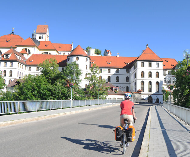 Das Bild zeigt eine Rennradfahrer von hinten. Er trägt einen Helm und ein rotes Trikot. Am Rad sind zwei Packtaschen befestigt. Der Radfahrer fährt über eine Brücke. Die Straße ist asphaltiert. Im Hintergrund ist der umfangreiche Gebäudekomplex des ehemaligen Benediktinerklosters St. Mang, mit seiner weißen Fassade, seinen unzähligen Fenstern und roten Dächern zu sehen. Der Himmel ist blau. Die Sonne scheint. 
