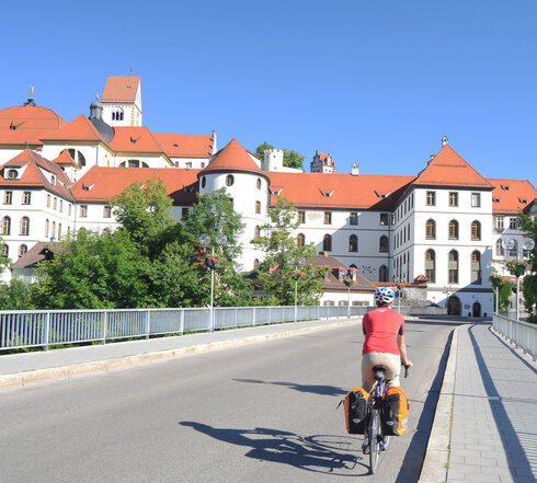 Das Bild zeigt eine Rennradfahrer von hinten. Er trägt einen Helm und ein rotes Trikot. Am Rad sind zwei Packtaschen befestigt. Der Radfahrer fährt über eine Brücke. Die Straße ist asphaltiert. Im Hintergrund ist der umfangreiche Gebäudekomplex des ehemaligen Benediktinerklosters St. Mang, mit seiner weißen Fassade, seinen unzähligen Fenstern und roten Dächern zu sehen. Der Himmel ist blau. Die Sonne scheint. 