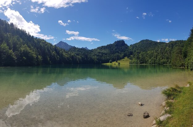 Die Aufnahme zeigt eine ruhige Szene am Ufer eines Sees - des Alatsees. Der lebhaftes blaue Alatsee liegt eingebettet in eine üppige Baumlandschaft. Die Wasseroberfläche ist ruhig und spiegelt die umgebenden Grüntöne wider. Auf der rechten Seite des Bildes ist ein sandiger Strand zu erkennen, der der kühlen Bläue des Sees etwas Wärme verleiht. Der Himmel darüber ist ein strahlendes Blau, gesprenkelt mit flauschigen weißen Wolken. In der Ferne erheben sich majestätische Berge, deren Gipfel fast bis zum Himmel reichen. 
