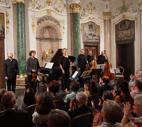 Das Bild zeigt eine Gruppe Musiker auf einer niedrigen Bühne stehen. Einige haben Geigen in den Händen. Auch ein Cello und ein Kontrabass ist dabei. Im Hintergrund steht ein Konzertflügel.  Die Wände des Raums sind prunkvoll verziert. Vor der Bühne sitzen zahlreiche Personen auf Stühlen der Bühne zugewandt und applaudieren. Der Raum ist hell erleuchtet. 