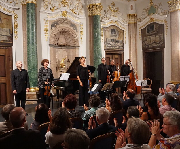 Das Bild zeigt eine Gruppe Musiker auf einer niedrigen Bühne stehen. Einige haben Geigen in den Händen. Auch ein Cello und ein Kontrabass ist dabei. Im Hintergrund steht ein Konzertflügel.  Die Wände des Raums sind prunkvoll verziert. Vor der Bühne sitzen zahlreiche Personen auf Stühlen der Bühne zugewandt und applaudieren. Der Raum ist hell erleuchtet. 