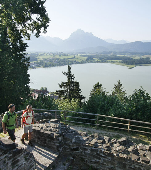 Das Bild zeigt eine friedvolle Szene von zwei Menschen, einem Mann und einer Frau, die in den alten Mauern einer verfallenen Burg herumgehen. Die Ruine befindet sich auf einer Anhöhe über dem Hopfensee und ist von einer lichten Berglandschaft und einem dichten Wald umgeben. Die Bergkette, majestätisch und hoch, bildet eine beeindruckende Kulisse für diese Szenerie. Der Himmel ist ein helles Blau, was zu der allgemeinen Ruhe des Bildes beiträgt. Die beiden Menschen, in legerer Kleidung, scheinen einen gemütlichen Spaziergang am Ufer zu genießen. Das Bild vermittelt ein Gefühl von Frieden und Ruhe und ist somit ein faszinierender Anblick.