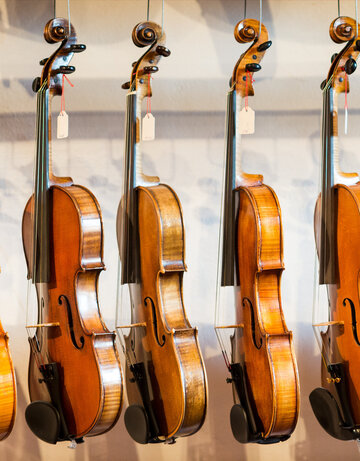Das Bild zeigt eine Sammlung von Geigen, die jeder einzeln von einem Haken an einer Wand hängt. Die Geigen sind in einer Reihe angeordnet, mit jedem Instrument gleich in eine Richtung zeigend. Die Geigen sind unterschiedlicher Größe, was darauf hindeutet, dass sie für verschiedene Altersgruppen oder Spielweisen konzipiert sind. Die Geigen sind aus Holz und haben eine reiche, warme Braunfärbung, die vor dem weißen Hintergrund im Bild gut zur Geltung kommt. Die Saiten der Geigen sind sichtbar und tragen dazu bei, die Gesamtästhetik des Bildes zu vervollständigen. Die Anordnung der Geigen und deren Positionierung an der Wand erzeugen ein Gefühl von Ordnung und Harmonie