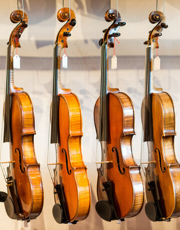 Das Bild zeigt eine Sammlung von Geigen, die jeder einzeln von einem Haken an einer Wand hängt. Die Geigen sind in einer Reihe angeordnet, mit jedem Instrument gleich in eine Richtung zeigend. Die Geigen sind unterschiedlicher Größe, was darauf hindeutet, dass sie für verschiedene Altersgruppen oder Spielweisen konzipiert sind. Die Geigen sind aus Holz und haben eine reiche, warme Braunfärbung, die vor dem weißen Hintergrund im Bild gut zur Geltung kommt. Die Saiten der Geigen sind sichtbar und tragen dazu bei, die Gesamtästhetik des Bildes zu vervollständigen. Die Anordnung der Geigen und deren Positionierung an der Wand erzeugen ein Gefühl von Ordnung und Harmonie