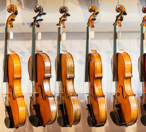 Das Bild zeigt eine Sammlung von Geigen, die jeder einzeln von einem Haken an einer Wand hängt. Die Geigen sind in einer Reihe angeordnet, mit jedem Instrument gleich in eine Richtung zeigend. Die Geigen sind unterschiedlicher Größe, was darauf hindeutet, dass sie für verschiedene Altersgruppen oder Spielweisen konzipiert sind. Die Geigen sind aus Holz und haben eine reiche, warme Braunfärbung, die vor dem weißen Hintergrund im Bild gut zur Geltung kommt. Die Saiten der Geigen sind sichtbar und tragen dazu bei, die Gesamtästhetik des Bildes zu vervollständigen. Die Anordnung der Geigen und deren Positionierung an der Wand erzeugen ein Gefühl von Ordnung und Harmonie