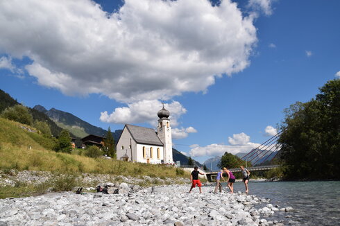 Das Bild zeigt vier Personen, die in einem Bachbett stehen. Das Ufer es Bachbetts ist mit groben Steinen und Kies bedeckt. Sie haben die Schuhe ausgezogen und treten ins Wassern. Im Hintergrund steht eine Kirche mit Kirchturm. Dahinter erheben sich die Berge. Der Himmel ist blau. Die Sonne scheint. 