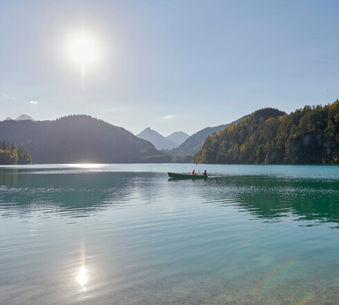 Das Bild fängt eine friedliche Szene eines Sees ein, der zwischen gebirgigen Landschaften eingebettet ist. Hier handelt es sich um den tiefblauen Alpsee, der sich bis zum Horizont ausdehnt, seine ruhige Oberfläche spiegelt die Umgebung wider. Zwei Personen sind dabei zu sehen, wie sie in der Mitte des Sees ein kleines Boot rudern, ihre Gestalten klein im Vergleich zur Weite des Wassers. Die Sonne scheint hell am Himmel, wodurch ein warmes Licht über die gesamte Szene gestreut wird und einen schönen Kontrast zur kühlen Blaufärbung des Sees schafft. Die Berge im Hintergrund, teilweise von den Strahlen der Sonne verdeckt, verleihen der Szene eine Tiefe und das Gefühl von Weite. 
