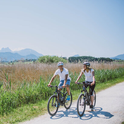 Das Bild zeigt einen Mann und eine Frau beim Radfahren auf einer Schotterstraße. Die Straße ist von üppigem grünem Gras und hohen Gräser umgeben. Die Radfahrer sind in der gleichen Richtung, was auf eine gemeinsame Reise oder ein Abenteuer schlägt. Die Kulisse des Bildes ist eine atemberaubende Aussicht von Bergen, die ein Gefühl von Abenteuer und Erkundung der Szene hinzufügt. Die Radfahrer sind in lässiger Kleidung, geeignet für einen Tag des Radfahrens. Die Gesamtatmosphäre des Bildes ist eine von Ruhe und Genuss der Natur.