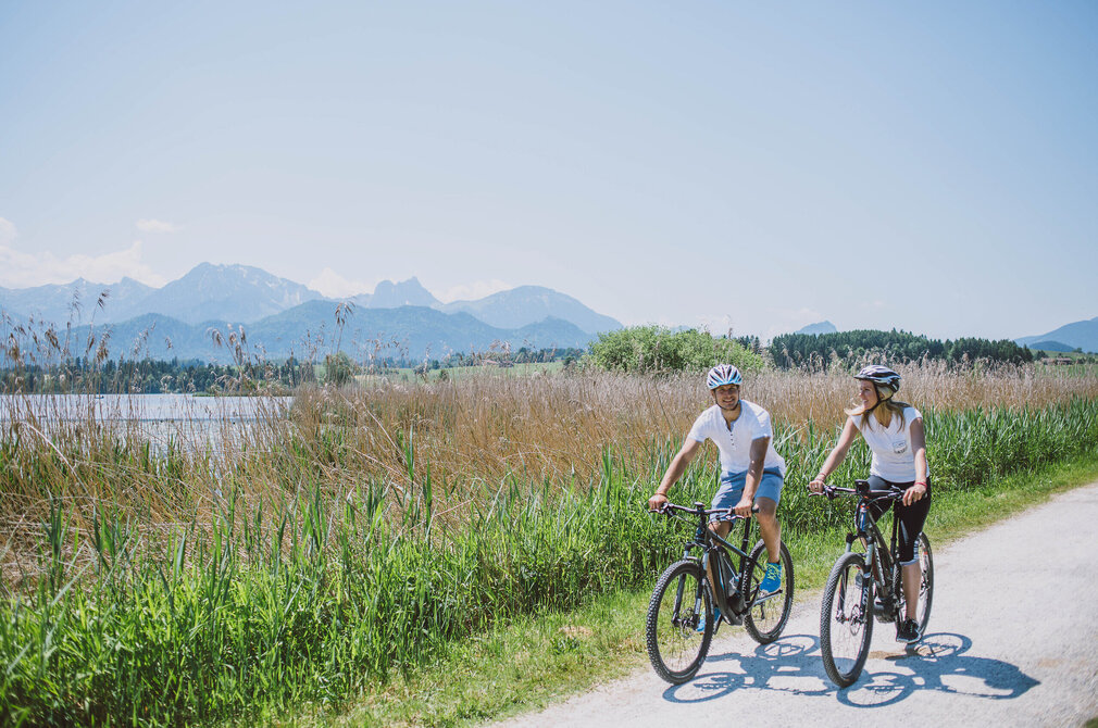 Das Bild zeigt einen Mann und eine Frau beim Radfahren auf einer Schotterstraße. Die Straße ist von üppigem grünem Gras und hohen Gräser umgeben. Die Radfahrer sind in der gleichen Richtung, was auf eine gemeinsame Reise oder ein Abenteuer schlägt. Die Kulisse des Bildes ist eine atemberaubende Aussicht von Bergen, die ein Gefühl von Abenteuer und Erkundung der Szene hinzufügt. Die Radfahrer sind in lässiger Kleidung, geeignet für einen Tag des Radfahrens. Die Gesamtatmosphäre des Bildes ist eine von Ruhe und Genuss der Natur.